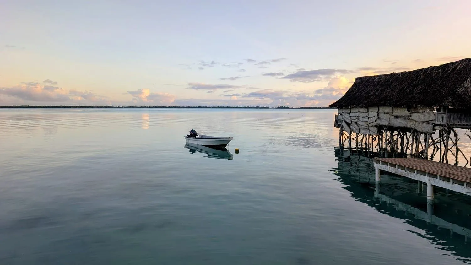 A moored boat on a still lagoon at dawn, with a thatched building standing on stilts over the water beside its jetty.