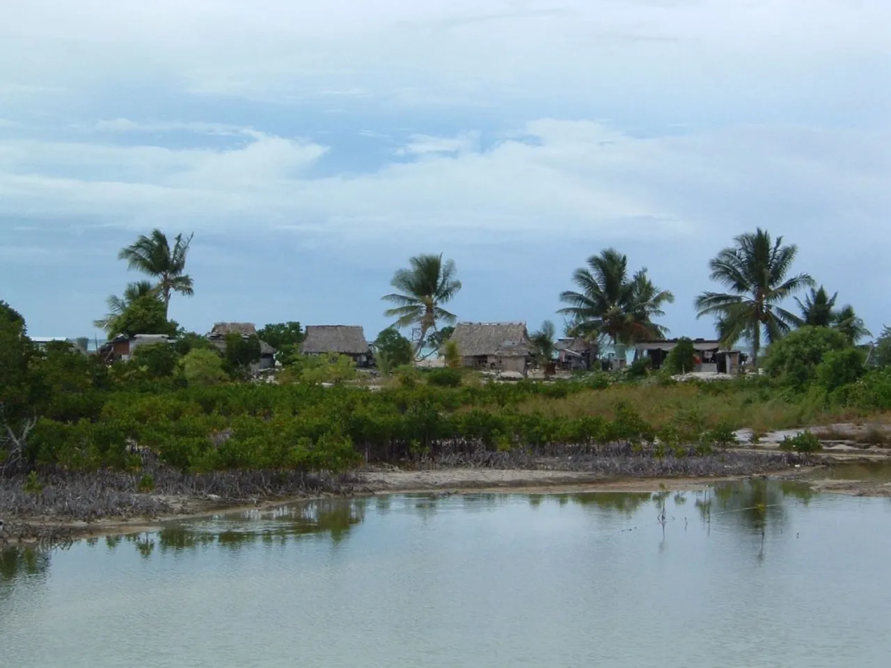 A village on the lagoon shore at Tarawa, with palms and thatched buildings behind the water.